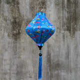 A Lozenge shaped Vietnamese lantern with a print of dragonflies, lady bugs and bees on bright blue background hanging in front of a grey wall.