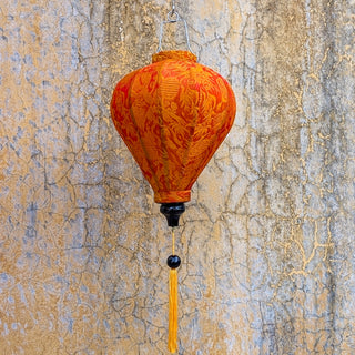 Balloon shaped Vietnamese lantern made with orange Jacquard silk fabric hanging in front of a cracked yellow and orange wall in Old Town Hoi An, Vietnam.