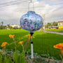 Round shaped silk lantern with koi fish, lotus flowers and bamboo trees hanging in a rural setting with green fields and flowers.