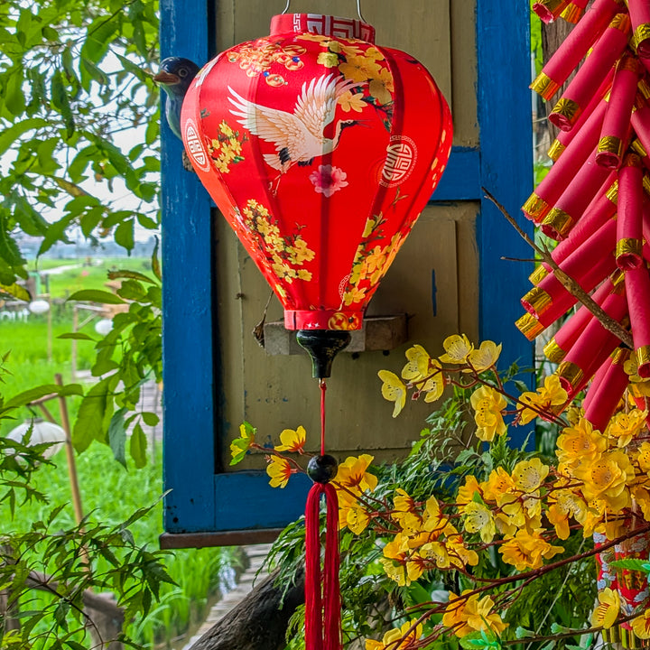 Red silk lantern with yellow and pink flowers and a bird design, hanging outdoors with greenery and firecrackers in the background.