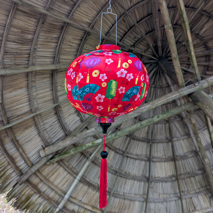Red silk lantern with pink flowers, gold coins, fans and lanterns hanging against a thatched roof background