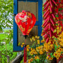 A vibrant red Hoi An silk lantern adorned with elegant white cranes, golden floral patterns, and traditional Vietnamese motifs. The lantern hangs near a blue wooden window, surrounded by lush green foliage, yellow apricot blossoms, and festive red firecrackers, creating a warm and festive atmosphere.
