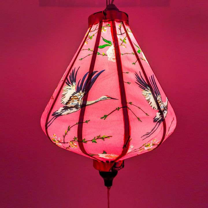 Glowing teardrop shaped lantern with print featuring large heron birds flying amongst pink flowers on a bright pink background.