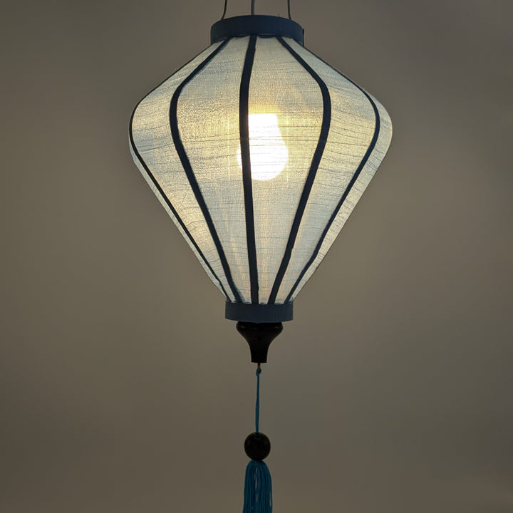 A hanging light blue polyester linen lantern with a glowing bulb inside, featuring a blue tassel at the bottom