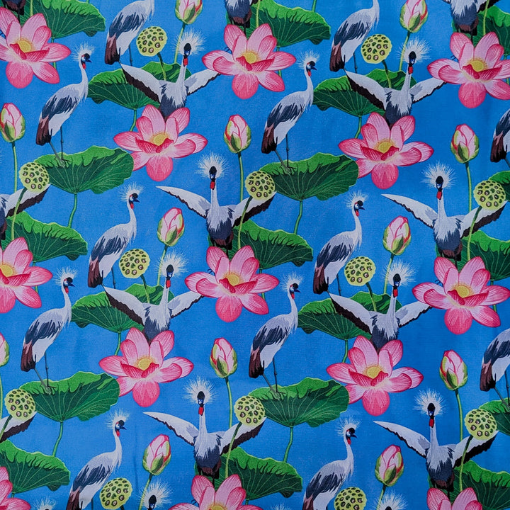 A striking fabric design featuring a vibrant blue background adorned with grey-crowned cranes standing among large green lotus leaves. The pattern includes blooming pink lotus flowers, unopened buds, and seed pods. The cranes, with their distinctive golden crests and black-and-white plumage.