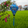 A vibrant blue Hoi An silk lantern featuring a delicate pattern of love birds perched among blossoming cherry branches. The intricate floral design is accentuated by the lantern’s elegant shape and a matching blue tassel. Suspended from a bougainvillea branch, the lantern contrasts beautifully with the lush green rice fields and traditional Vietnamese village houses in the background.