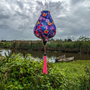 A Garlic shaped lantern with pink tassel hanging in front of boats in a river and green foliage. The lantern has a print of vibrant and colourful array of hibiscus flowers in various shades of colours. 