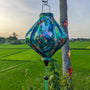 Lozenge shape Vietnamese silk lantern hanging in a lush green rice field featuring a print of parrots.