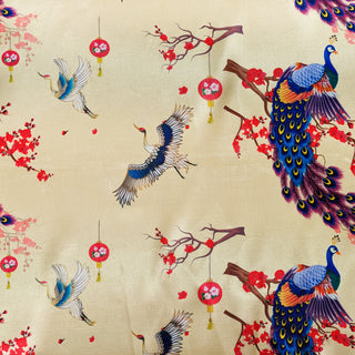 A closeup of polyester silk fabric with crane birds in flight, large flamboyant peacocks, dainty lanterns hanging on branches with pink and red flower blossoms on a cream background.