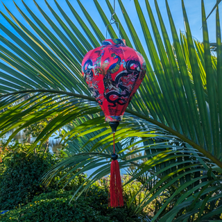 Balloon shaped lantern with red tassel adorned with intricate prints of dragons, peacocks, and butterflies against a striking red background set in front of a river with large palm leaves.