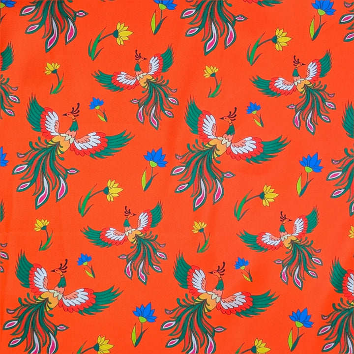 Close up of polyester and silk fabric featuring phoenix birds with green orange and white wings in flight amongst yellow and blue flowers on a bright orange background.