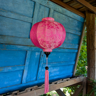 Pink Jacquard silk round lantern with tassel hanging in front of a blue wooden wall in a garden setting
