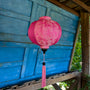 Pink Jacquard silk round lantern with tassel hanging in front of a blue wooden wall in a garden setting