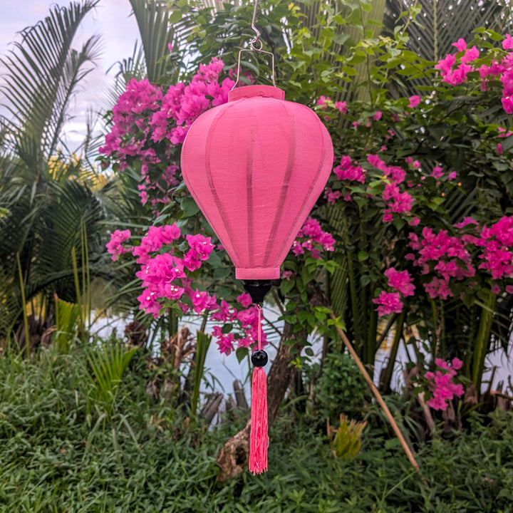 A pink poly linen fabric Hoi An Lantern hanging with a matching tassel suspended outdoors among green foliage and bright pink bougainvillea flowers.