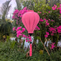 A pink poly linen fabric Hoi An Lantern hanging with a matching tassel suspended outdoors among green foliage and bright pink bougainvillea flowers.