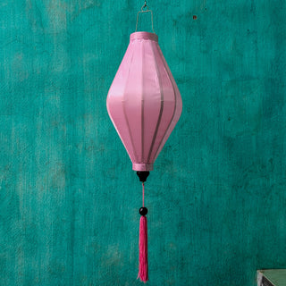 Tube shaped Vietnamese lantern in light pink plain silk with a bright pink tassel hanging in front of a bright blue wall.