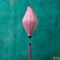 Tube shaped Vietnamese lantern in light pink plain silk with a bright pink tassel hanging in front of a bright blue wall.