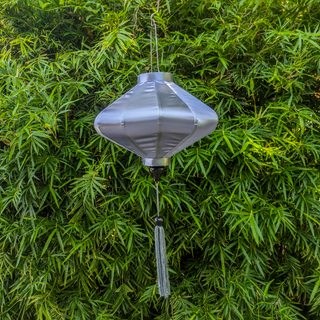 UFO shaped Vietnamese lantern made with steel, bamboo and grey polyester silk fabric hanging in front of a lush green plant.