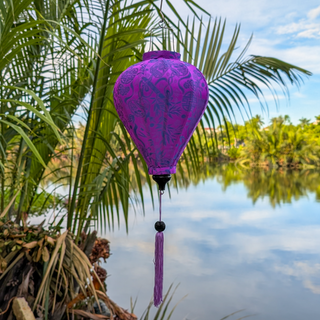 Balloon shaped Jacquard silk Purple lantern with matching purple tassel hanging by a river in Hoi An with palm trees in the background