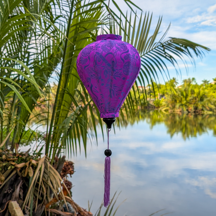 Balloon shaped Jacquard silk Purple lantern with matching purple tassel hanging by a river in Hoi An with palm trees in the background