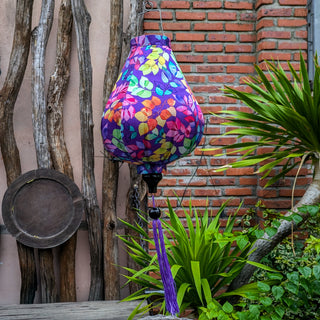 Purple silk lantern with colourful floral-pattern hanging  in front of a brick wall with plants and wooden branches.