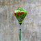 A striking green Hoi An silk lantern adorned with red folding fans, delicate plum blossoms, and traditional circular motifs. The lantern's elegant shape and rich colors stand out against a textured, weathered wall background. A matching green tassel hangs from the base, adding to its charm and cultural elegance.