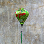 A striking green Hoi An silk lantern adorned with red folding fans, delicate plum blossoms, and traditional circular motifs. The lantern's elegant shape and rich colors stand out against a textured, weathered wall background. A matching green tassel hangs from the base, adding to its charm and cultural elegance.