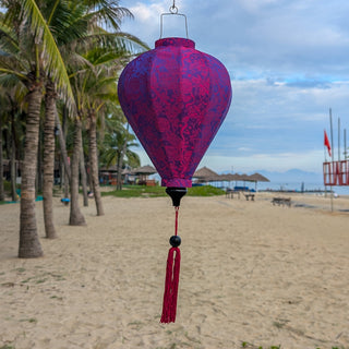 Purple jacquard silk lantern with tassel hanging on Cua Dai Beach with palm trees and umbrellas in the background.