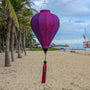 Purple jacquard silk lantern with tassel hanging on Cua Dai Beach with palm trees and umbrellas in the background.