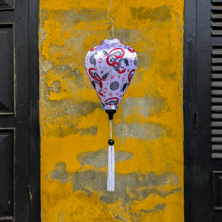 A balloon shaped lantern featuring entwined red serpentines and black dragons surrounded by lanterns, symbols, hanging in front of a yellow wall with black doors on either side.