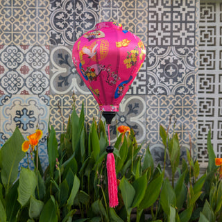 Pink decorative lantern with floral patterns hanging in front of a tiled wall with flowers blooming below.