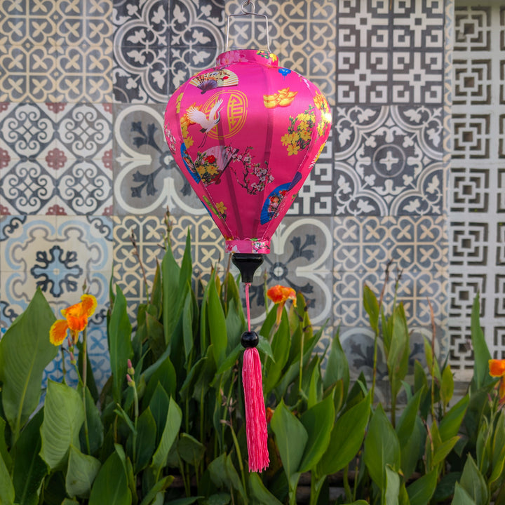 Pink decorative lantern with floral patterns hanging in front of a tiled wall with flowers blooming below.