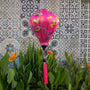 Pink decorative lantern with floral patterns hanging in front of a tiled wall with flowers blooming below.