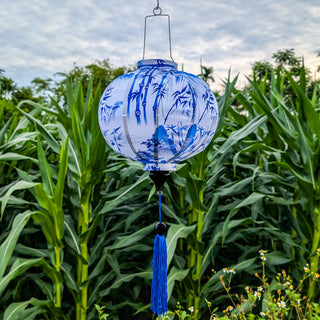 A round shape Vietnamese lantern with a blue print of bamboo and temples against white silk hanging in front of a corn field in Hoi An, Vietnam.
