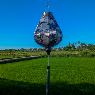 Garlic shaped lanterns featuring a print of mountains, trees, temples, men in boats, and large rocks. A beautiful palette of light pinks and purples against a serene creamy background hanging in front of lush green rice fields and blue open skies.