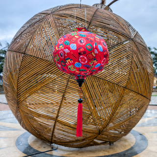 Red round lantern with colourful Tet / Lunar New Year patterns hanging in front of a woven spherical structure.