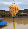Yellow lantern with floral patterns hanging over the Thu Bon River with traditional Hoi An buildings in the background.