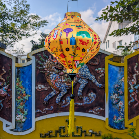 Lozenge shaped yellow lantern with colourful Lunar New Year motif's hanging in front of a temple in the background