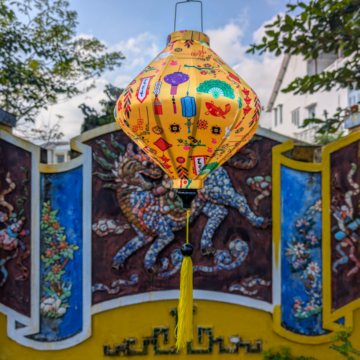 Lozenge shaped yellow lantern with colourful Lunar New Year motif's hanging in front of a temple in the background