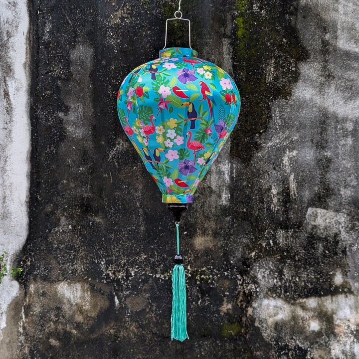 Balloon shaped lantern with colourful print featuring tropical birds and plants on light blue backdrop hanging with a tassel against a textured moss covered wall.