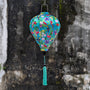 Balloon shaped lantern with colourful print featuring tropical birds and plants on light blue backdrop hanging with a tassel against a textured moss covered wall.