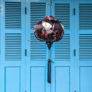 Round shaped Vietnamese Hoi An lantern featuring red-headed white cranes amongst pink cherry blossoms against a black background hanging in front of blue shutters. Perfect for home decor.