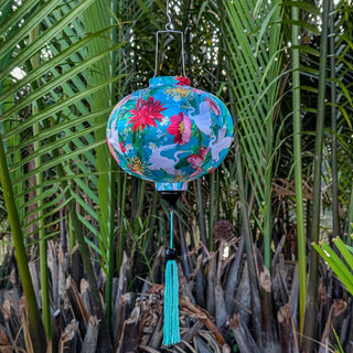 Round shape Vietnamese silk lantern hanging in front of palm leaves featuring vibrant cranes & floral print surrounded by a lush flowers.
