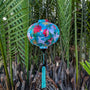 Round shape Vietnamese silk lantern hanging in front of palm leaves featuring vibrant cranes & floral print surrounded by a lush flowers.