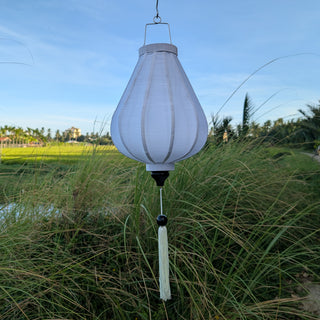 Garlic shaped white poly linen fabric lantern with white tassel hanging suspended above reeds with sun rising on rice fields in the background