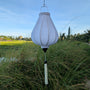 Garlic shaped white poly linen fabric lantern with white tassel hanging suspended above reeds with sun rising on rice fields in the background