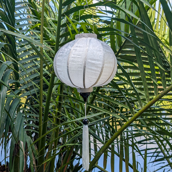 Hanging white Jacquard silk Vietnamese lantern with tassel against a green leafy background