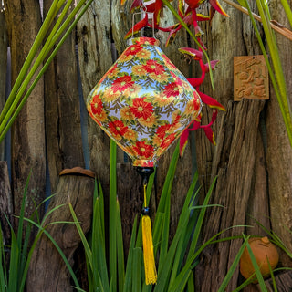 Lozenge shaped lantern with yellow and orange daisies hanging among plants and wooden planks in a garden