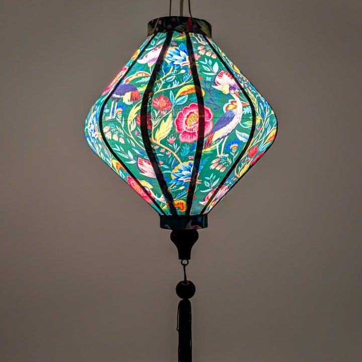 Glowing Lozenge shaped lantern with green tassel featuring a print of lush, tropical-inspired design with birds and flowers on a deep green silk fabric.