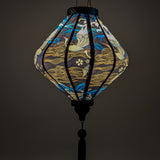 Glowing Lozenge shaped lantern featuring a print of birds in graceful flight, set against a sleek black background adorned with rolling waves, floating flowers and intricate gold illustrations.